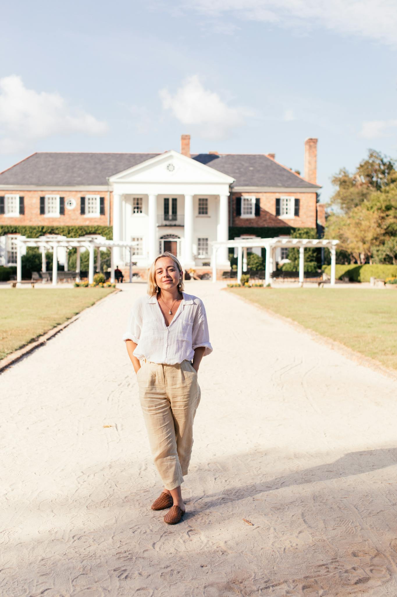 Woman walking on path leading to colonial-style mansion, exuding elegance and grace.